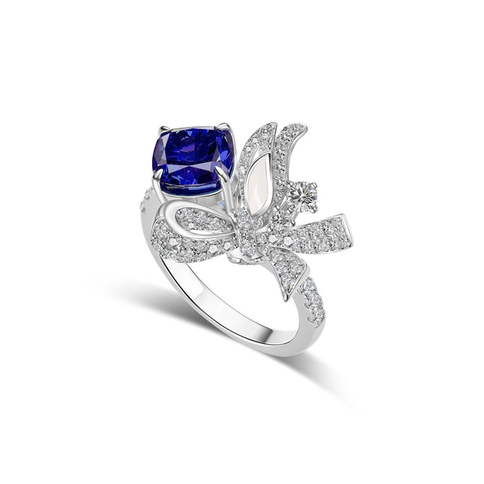 The Gem Vivi Blue Sapphire Drop Ribbon Accent Ring showcases a large cushion-cut sapphire in 18k white gold, highlighted by a delicate ribbon-inspired design adorned with sparkling stones.