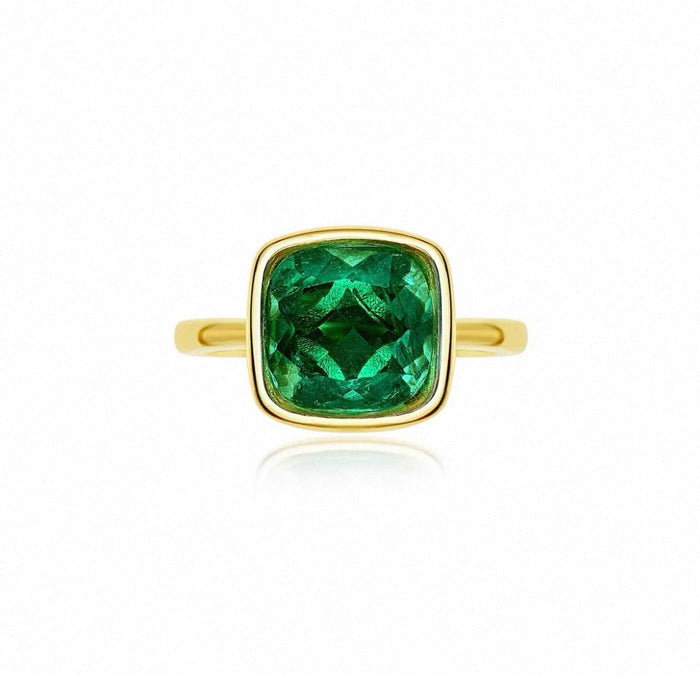 The Gem Vivi Chole Duo Glow Cushion Ring showcases a cushion cut emerald centerpiece set in an 18k yellow gold bezel, displayed on a plain white background.