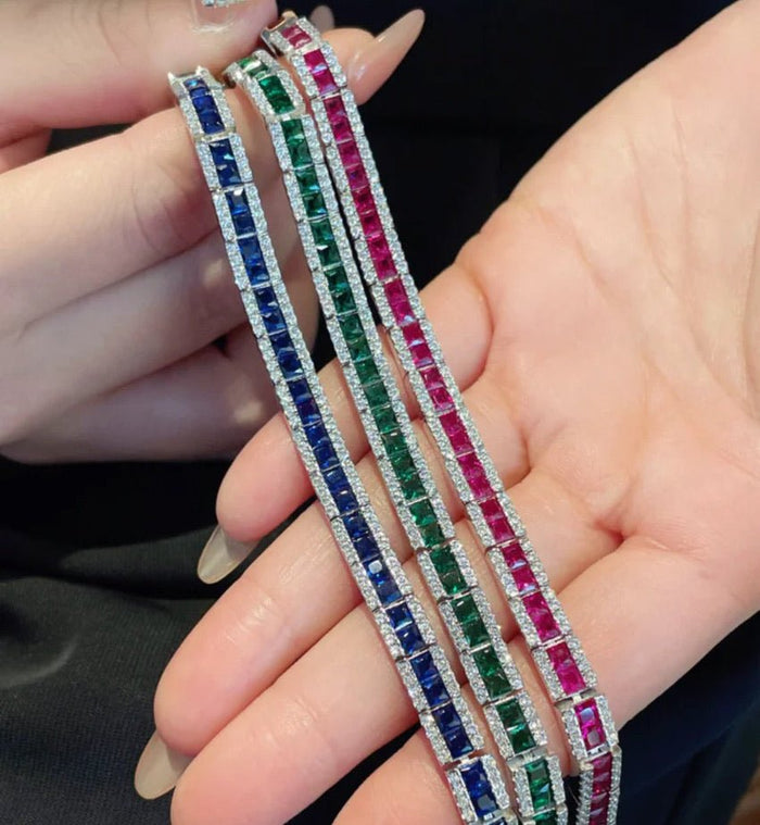 A hand holds three Gem Vivi Baguette Tennis Bracelets, each featuring square-cut lab created gemstones in blue, green, and pink, bordered by small clear stones.
