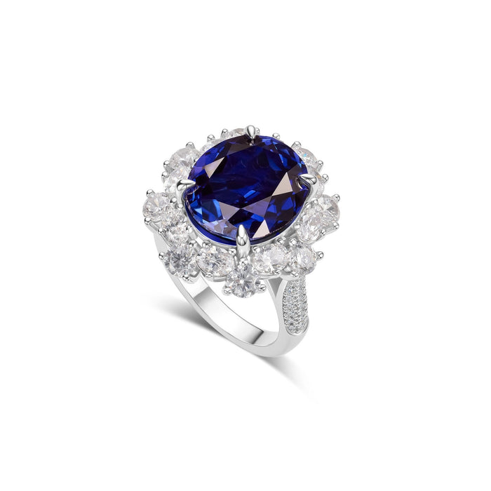 The Gem Vivi Blue Sapphire Large Halo Ring showcases a prominent blue gemstone in an oval setting, encircled by smaller clear stones on a diamond-accented band.