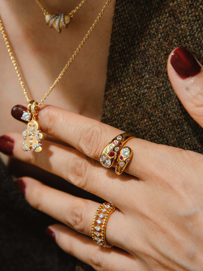 A close-up of a person wearing the Gem Vivi Pomegranate Red Wrap Ring in 18k yellow gold with cubic zirconia, paired with a gold chain necklace and dark red nail polish.