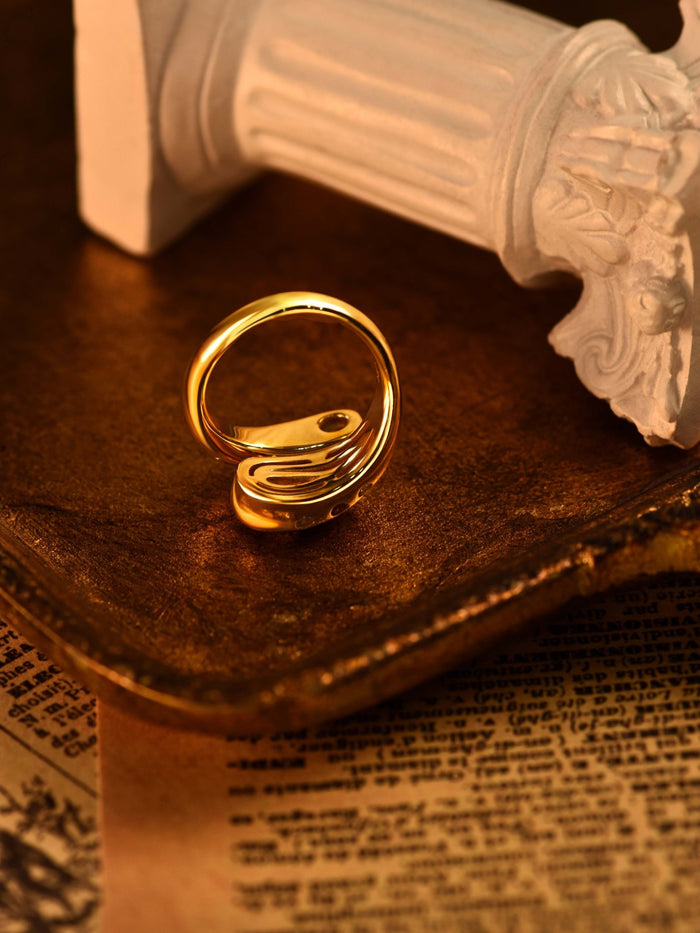 The Pomegranate Red Wrap Ring by Gem Vivi, featuring a modern abstract design in 18k yellow gold, is displayed on a bronze tray beside a broken white column and vintage newspaper clippings.