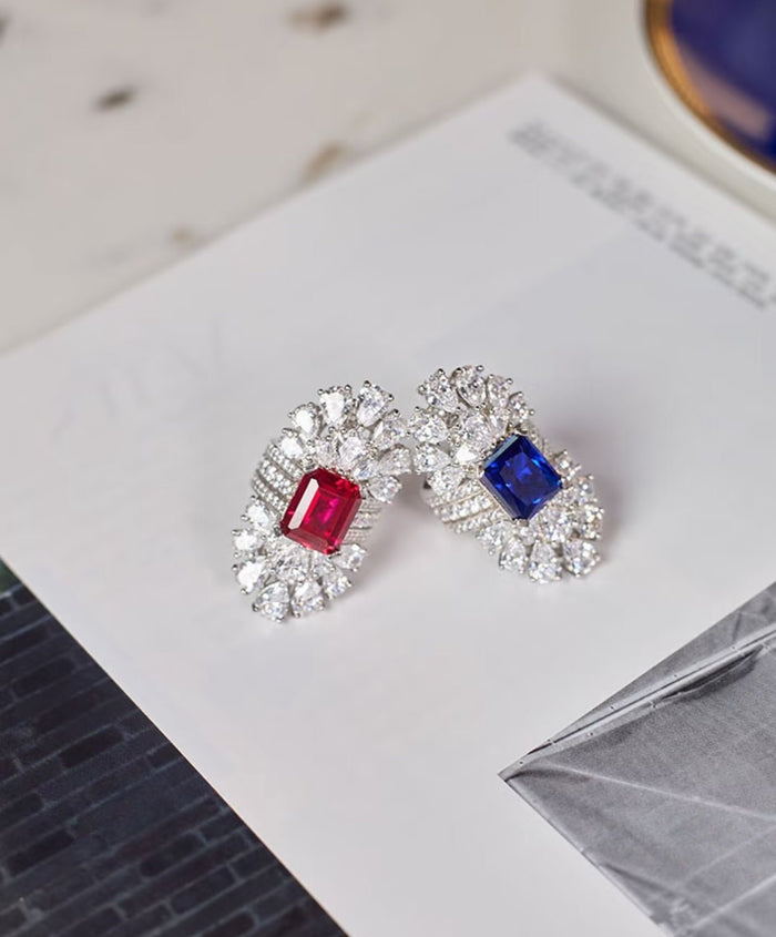 A pair of ornate earrings with lab-created red and blue gemstones framed by sparkling stones on an open book—an elegant match for your Gem Vivi Blue Radiant Cushion-Cut Sapphire Halo Ring or 18k white gold jewelry.