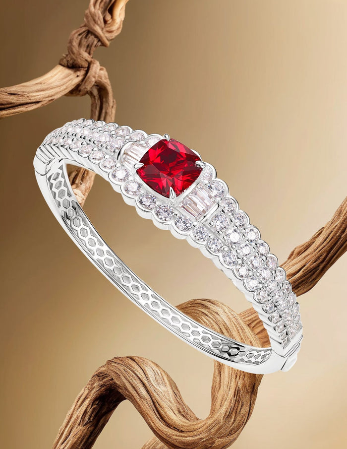 The Clara Square Ruby Bangle Bracelet by Gem Vivi features a square ruby centerpiece with sparkling accents, shown against a beige background with twisted wood details.