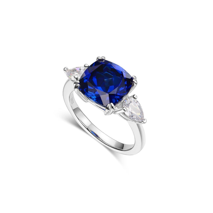 The Gem Vivi Blue Sapphire Trilogy Ring showcases a brilliant lab-created cushion cut sapphire in 18k white gold, accented by two pear-shaped clear stones.
