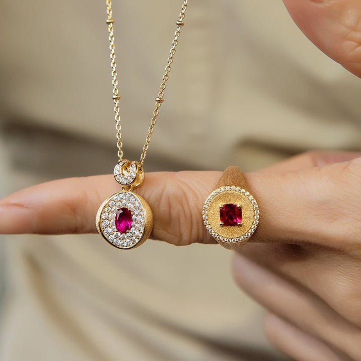 A hand displays the Gem Vivi Ivy Signet Ring in 18K yellow gold, set with a cushion-cut red gemstone and surrounded by small clear stones.