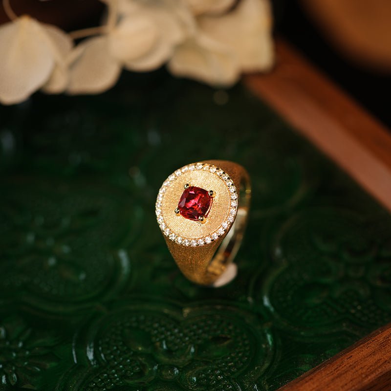 The Ivy Signet Ring by Gem Vivi features 18K yellow gold, a square red gemstone, and small white stones, elegantly displayed on an ornate dark green surface with a blurred floral background.