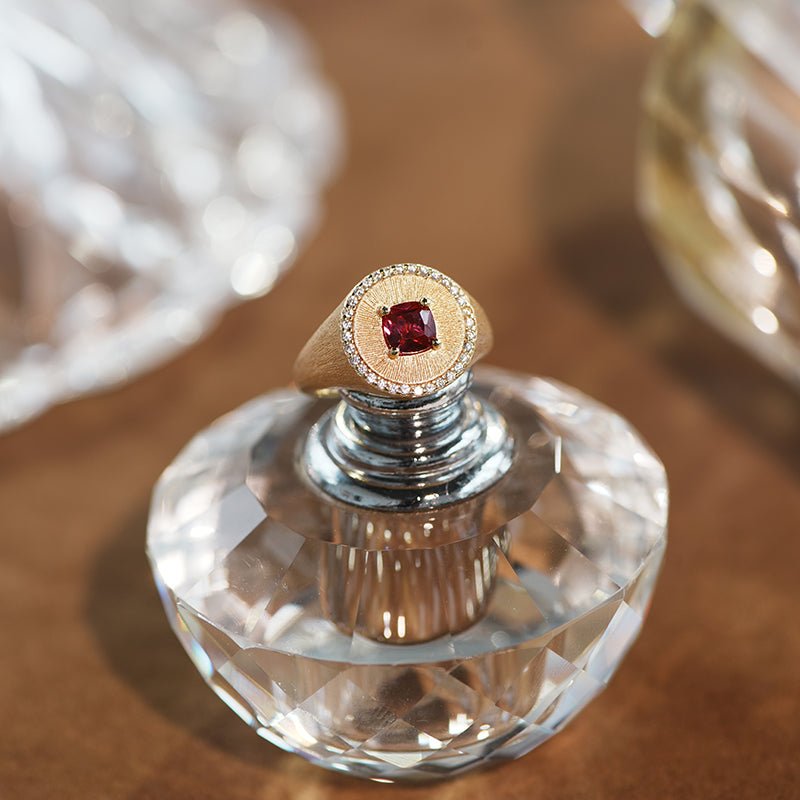The Ivy Signet Ring by Gem Vivi features a cushion-cut red stone set in 18K yellow gold with a halo of diamonds, beautifully displayed on a faceted glass perfume bottle.