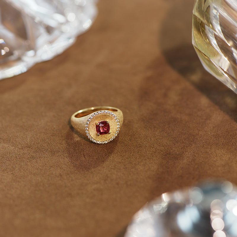 The Ivy Signet Ring by Gem Vivi features a cushion-cut red stone set in 18K yellow gold, accented with clear stones, and is displayed on a brown surface near glass objects.
