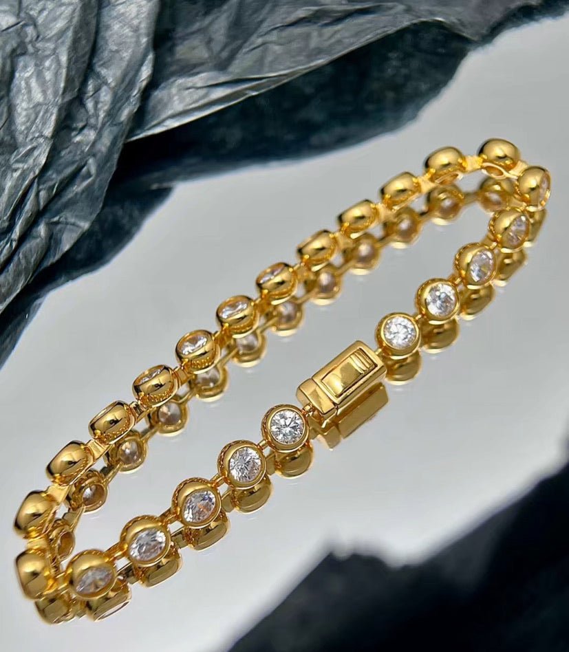 The Radiance Bezel Bracelet by Gem Vivi, crafted in 18K yellow gold with round cubic zirconia gemstones, shines elegantly on a reflective surface set against a textured background.