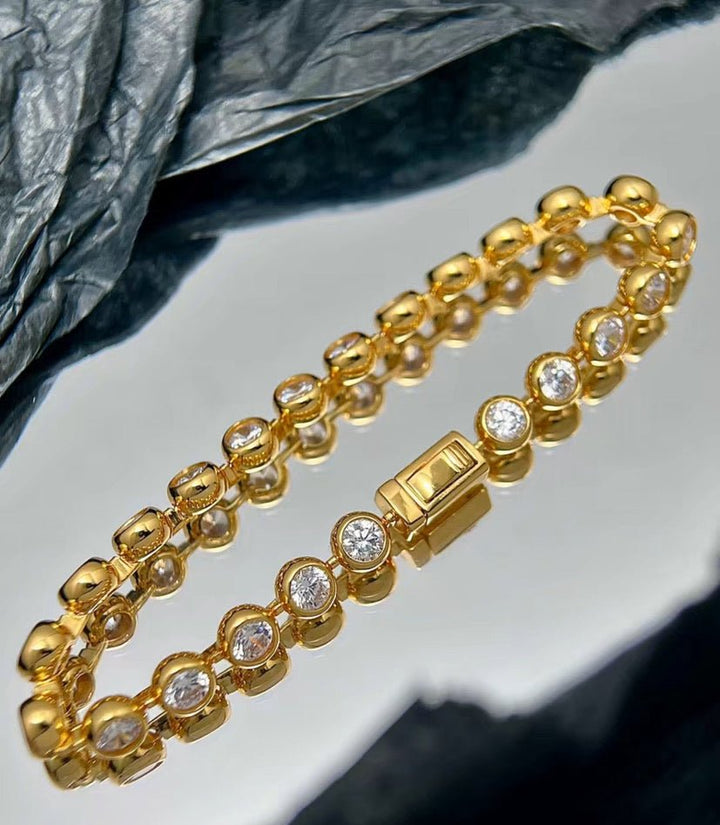 The Radiance Bezel Bracelet by Gem Vivi, crafted in 18K yellow gold with round cubic zirconia gemstones, shines elegantly on a reflective surface set against a textured background.