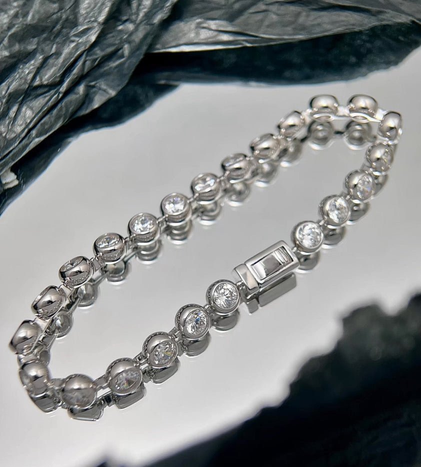 The Gem Vivi Radiance Bezel Bracelet, featuring round clear cubic zirconia stones in a silver setting, is displayed on a reflective surface against a backdrop of dark textured fabric.