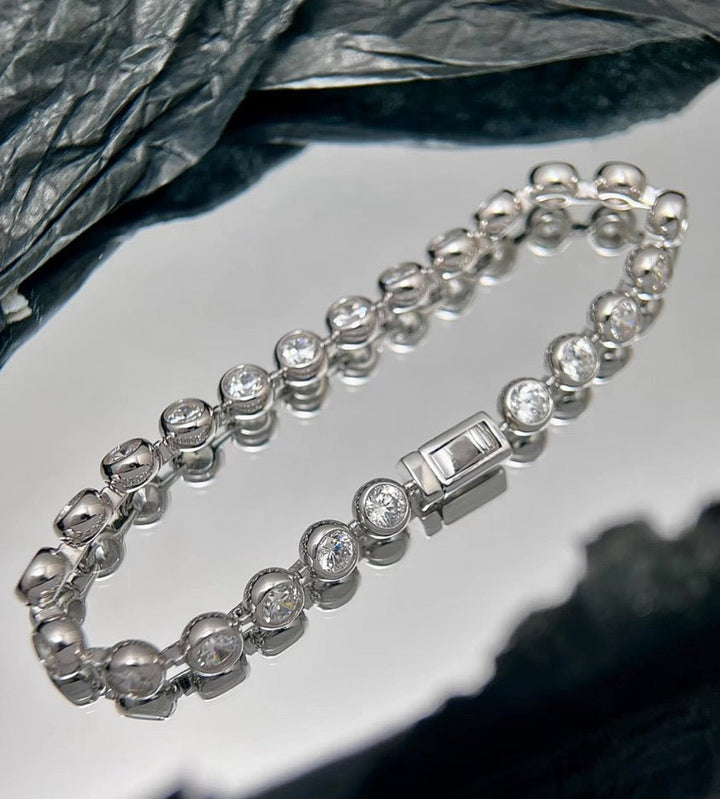 The Gem Vivi Radiance Bezel Bracelet, featuring round clear cubic zirconia stones in a silver setting, is displayed on a reflective surface against a backdrop of dark textured fabric.