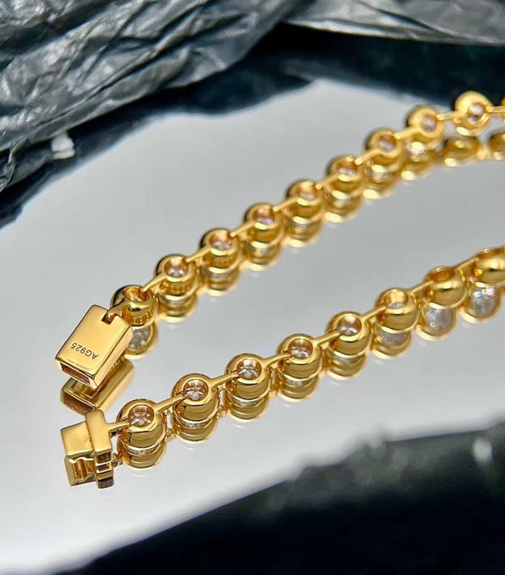 The Radiance Bezel Bracelet by Gem Vivi, crafted in 18K yellow gold and set with clear round cubic zirconia in each link, features a "925" marked clasp and is displayed on a reflective surface.