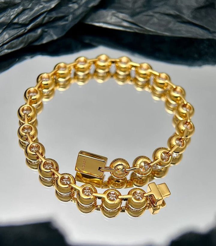 The Radiance Bezel Bracelet by Gem Vivi is crafted from 18K yellow gold, showcasing round links set with sparkling cubic zirconia, displayed on a reflective surface against a black textured background.