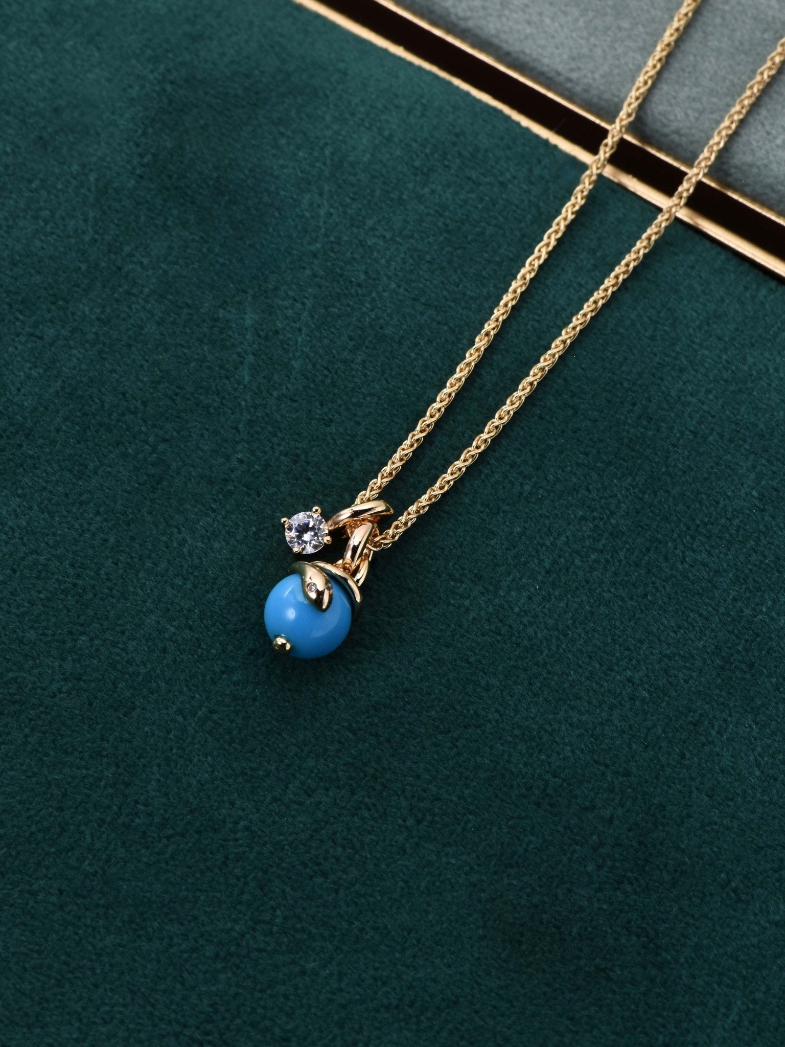The Serpentine Charm Necklace by Gem Vivi features 18K yellow gold with a blue charm and clear gemstone pendant, shown on green velvet beside a book.