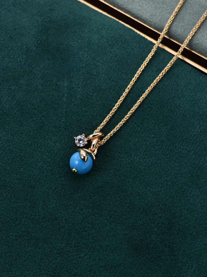 The Serpentine Charm Necklace by Gem Vivi features 18K yellow gold with a blue charm and clear gemstone pendant, shown on green velvet beside a book.