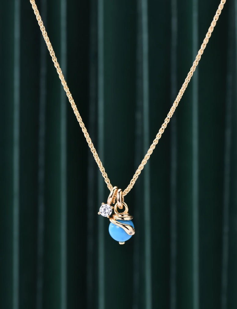 The Serpentine Charm Necklace by Gem Vivi features a blue charm with a round shell pearl pendant, an 18K yellow gold wrap accent, and a clear gemstone charm, shown on a dark green striped background.