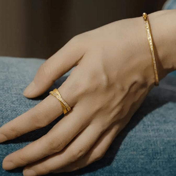 A hand rests on a leg, wearing a gold ring with a geometric design and Gem Vivi’s Hinged Baguette Glow Bangle Bracelet in 18K yellow gold, adorned with sparkling cubic zirconia.
