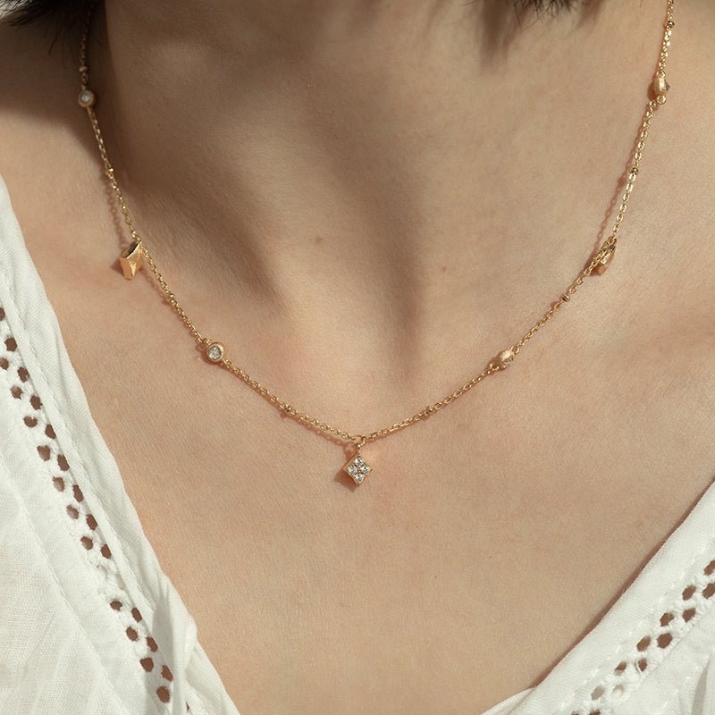 A person wearing the Gem Vivi Ivy Charm Station Necklace in 18K yellow gold, featuring small geometric charms, paired with a textured white V-neck top.
