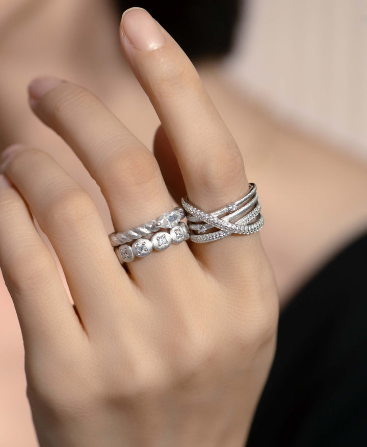 A hand adorned with multiple silver rings, including the Gem Vivi Sparkle Celeste Crossing Band on the ring and middle fingers, set against a neutral background.