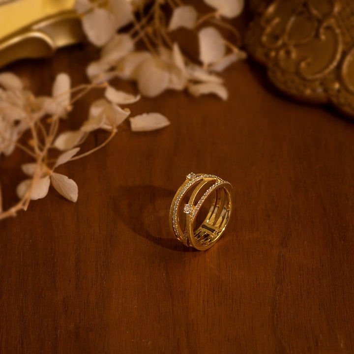 The Sparkle Celeste Crossing Band by Gem Vivi features intersecting 18K yellow gold bands with small cubic zirconia, displayed on wood with dried leaves and decorative accents in the background.