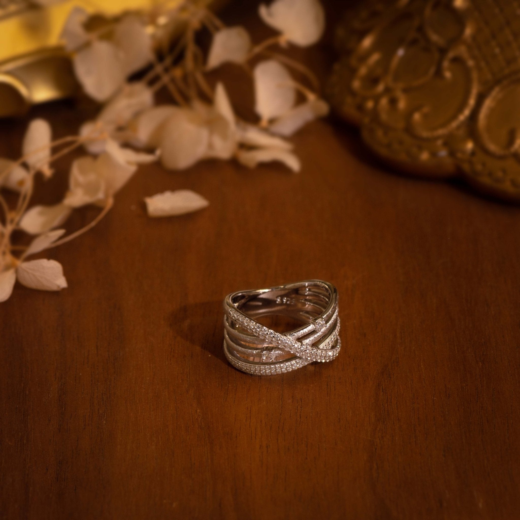 The Gem Vivi Sparkle Celeste Crossing Band features intertwined silver bands adorned with small Cubic Zirconia stones, displayed on a polished wooden surface with decorative objects and dried flowers.