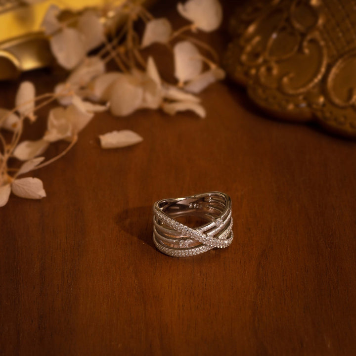 The Gem Vivi Sparkle Celeste Crossing Band features intertwined silver bands adorned with small Cubic Zirconia stones, displayed on a polished wooden surface with decorative objects and dried flowers.