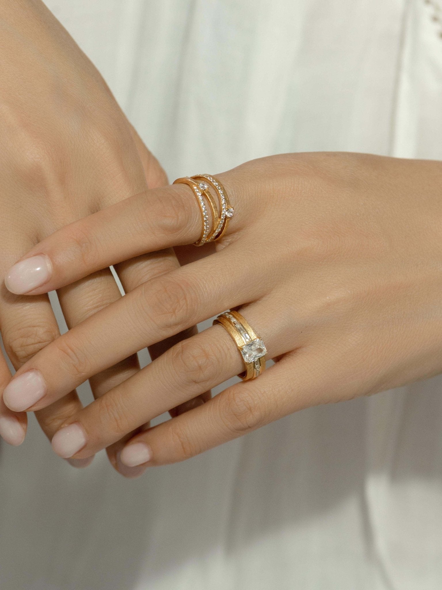 Close-up of hands wearing multiple gold rings, including Gem Vivi's Sparkle Celeste Crossing Band and a diamond ring on the left ring finger, all set in 18K yellow gold against a white fabric background.