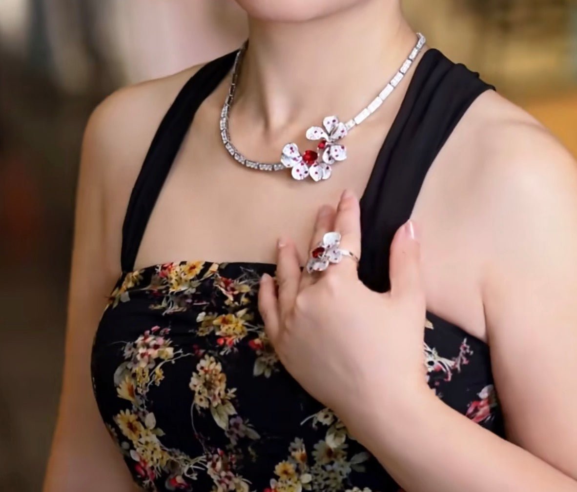 A woman in a sleeveless floral dress showcases a matching floral necklace and ring with white and red accents, featuring the elegant Crimson Petal Blossom Tennis Necklace by Gem Vivi.