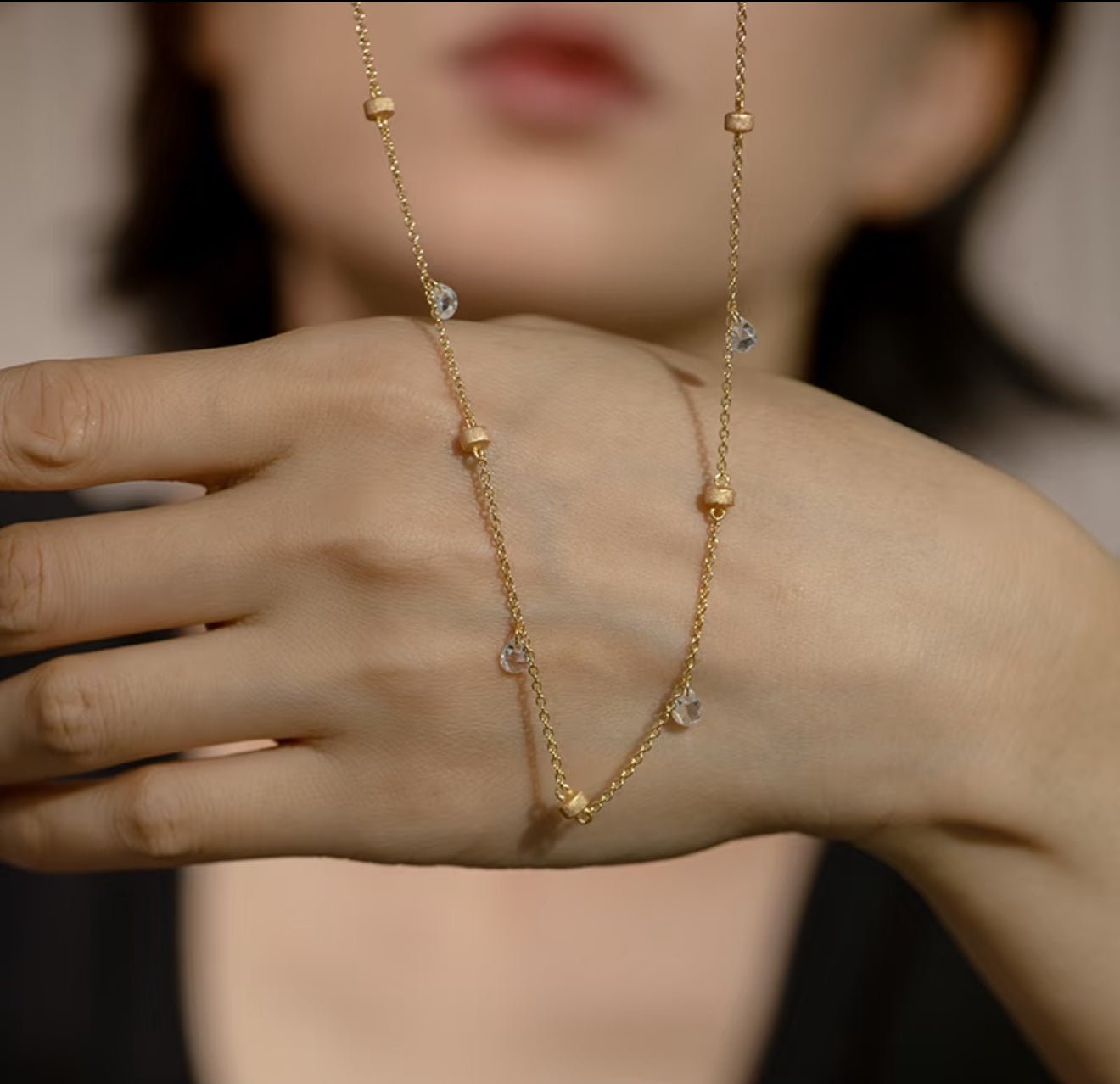The Raindrop Gold Necklace by Gem Vivi features small beads and sparkling cubic zirconia, shown draped over a hand with a blurred face in the background.