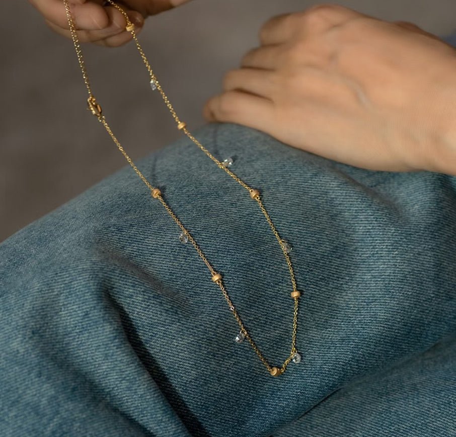 A hand holds the Raindrop Gold Necklace by Gem Vivi, featuring delicate 18K gold with small beads and sparkling cubic zirconia, resting on blue denim fabric.