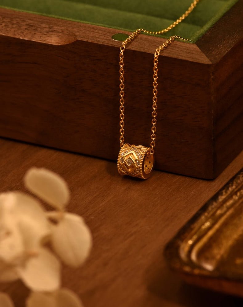 The Gem Vivi Lattice Barrel Necklace, featuring a gold chain and diamond lattice barrel pendant in 18K yellow gold, is displayed on wood near a green-lined box and a white flower.