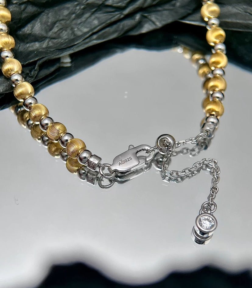 Close-up of the Gem Vivi Glow Dual Tone Bead Bracelet, featuring alternating 18K Yellow Gold and 925 Sterling Silver beads, a lobster clasp, extension chain, and small gemstone pendant, displayed on a reflective surface.
