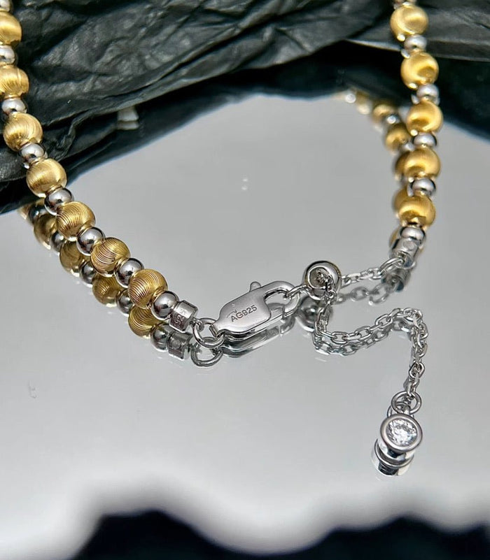 Close-up of the Gem Vivi Glow Dual Tone Bead Bracelet, featuring alternating 18K Yellow Gold and 925 Sterling Silver beads, a lobster clasp, extension chain, and small gemstone pendant, displayed on a reflective surface.