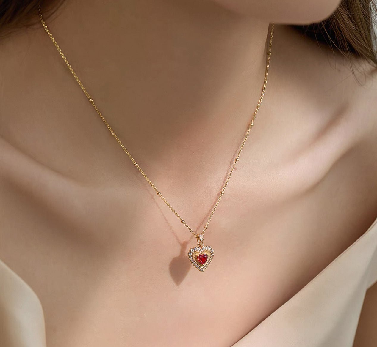 A woman wears the Gem Vivi Heart Ember Halo Necklace, featuring an 18K yellow gold heart-shaped pendant with a red center stone and sparkling cubic zirconia accents.