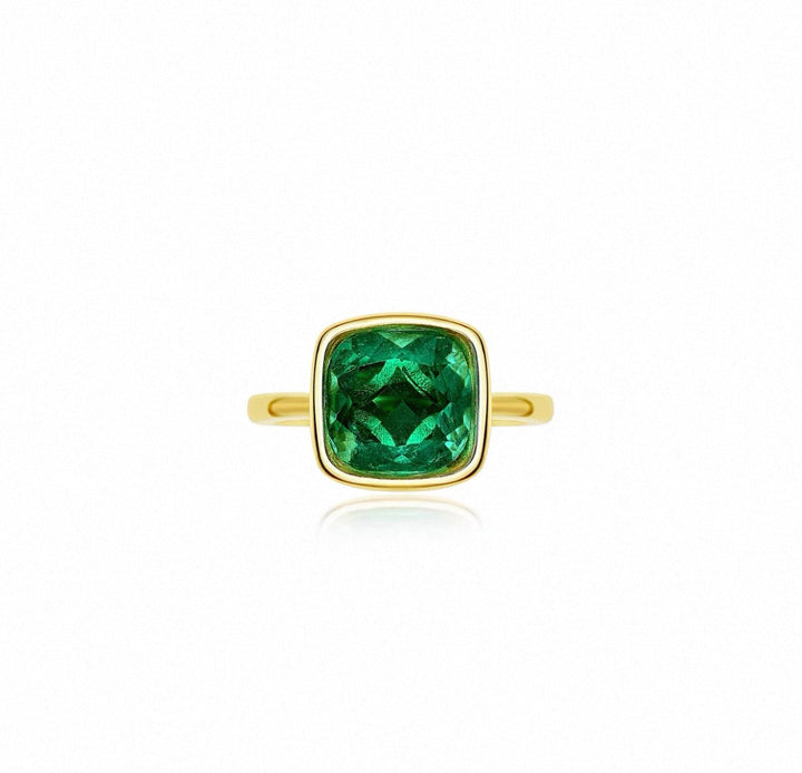 The Gem Vivi Chole Duo Glow Cushion Ring showcases a cushion cut emerald centerpiece set in an 18k yellow gold bezel, displayed on a plain white background.