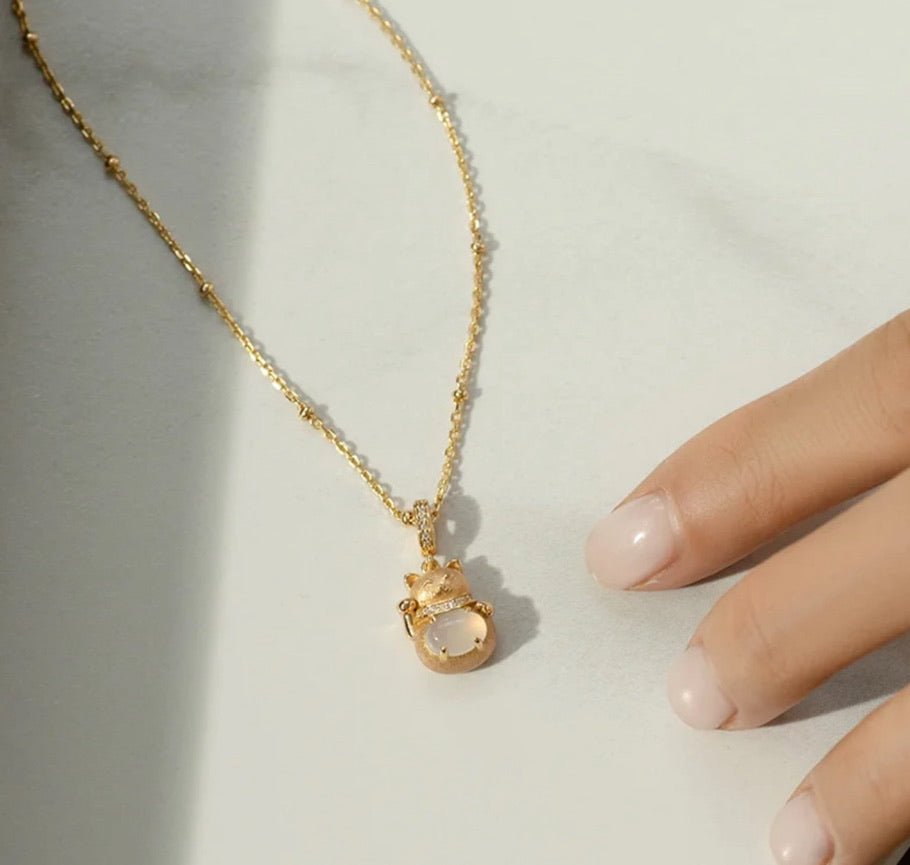 The Gem Vivi White Fortune Cat Necklace, featuring a gold chain and white stone pendant, is displayed on a light surface beside a hand with light pink manicured nails.