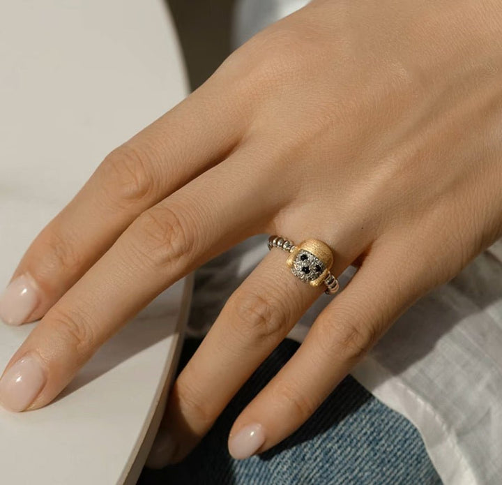 A hand with a natural manicure wears the Gem Vivi Dog In Golden Hat Ring, featuring 18K yellow gold and silver details. The person is resting their hand on a light-colored surface.