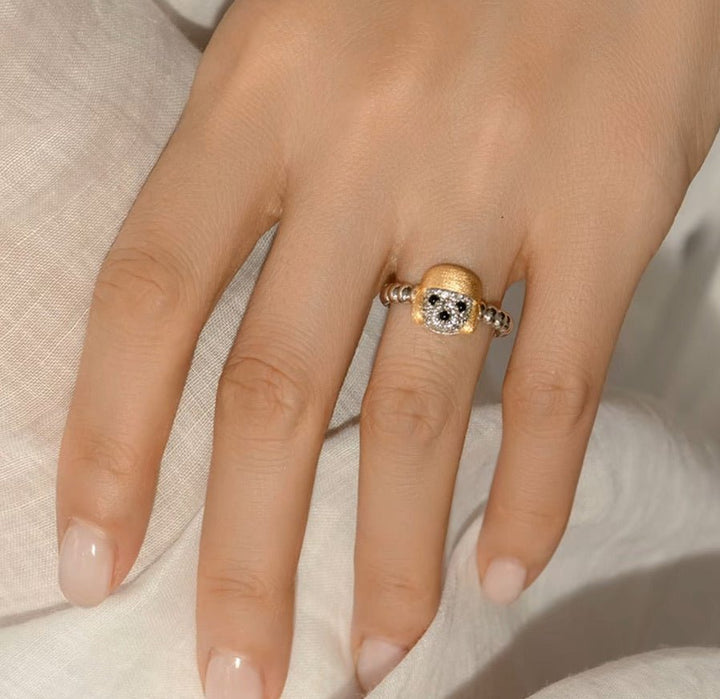 A hand with neatly manicured nails wears the Gem Vivi Dog In Golden Hat Ring, crafted in 18K Yellow Gold with a central diamond and decorative accents, set against a light fabric background.