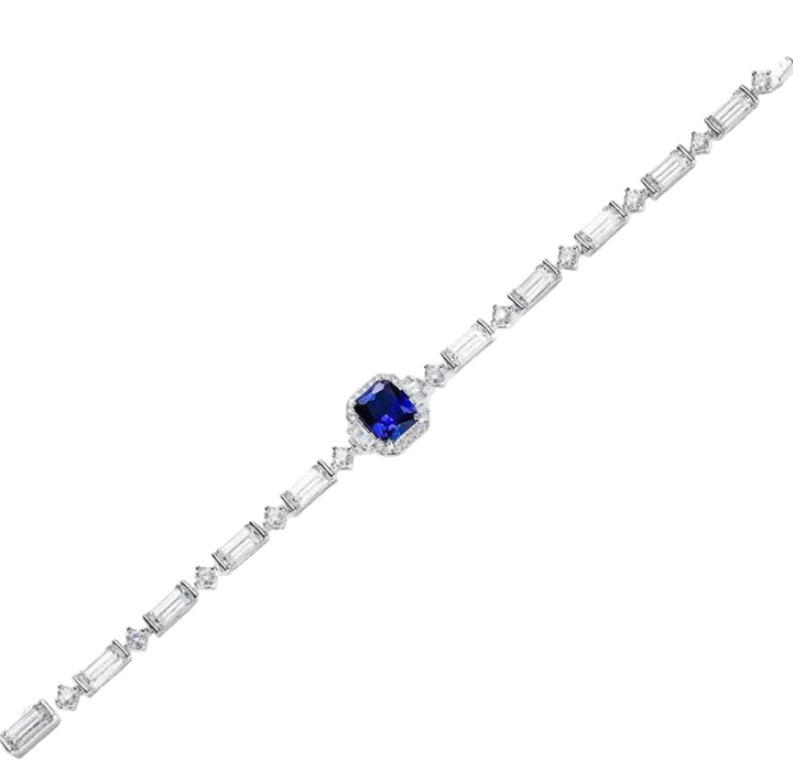 The Gem Vivi Blue Midnight Cascade Tennis Bracelet showcases rectangular clear stones and an emerald cut sapphire centerpiece, all set in radiant 18k white gold, displayed against a white background.