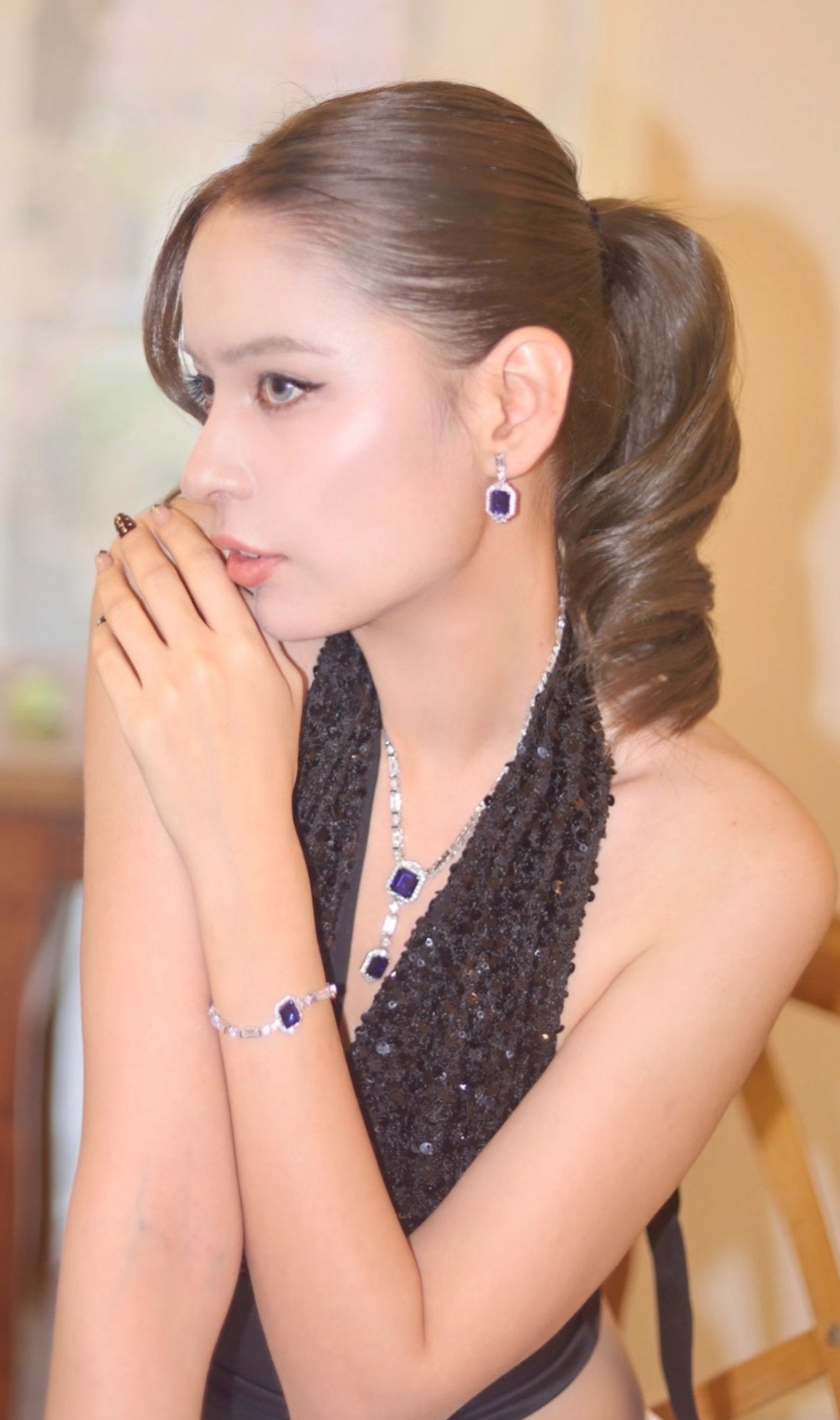 A woman with styled brown hair in a ponytail wears a black halter dress, blue gemstone jewelry—emerald-cut sapphire earrings, a necklace, and the Gem Vivi Blue Midnight Cascade Tennis Bracelet in 18k white gold.