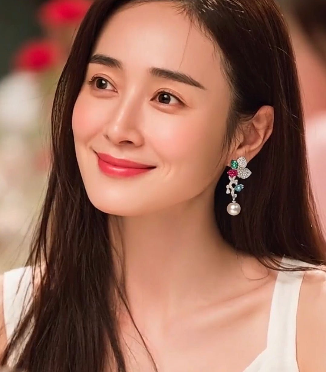 A woman with long dark hair smiles, wearing a white sleeveless top and Gem Vivi’s Pearl Glow Drop Earrings, expertly crafted from 18k White Gold and adorned with an Australian White Shell Pearl.