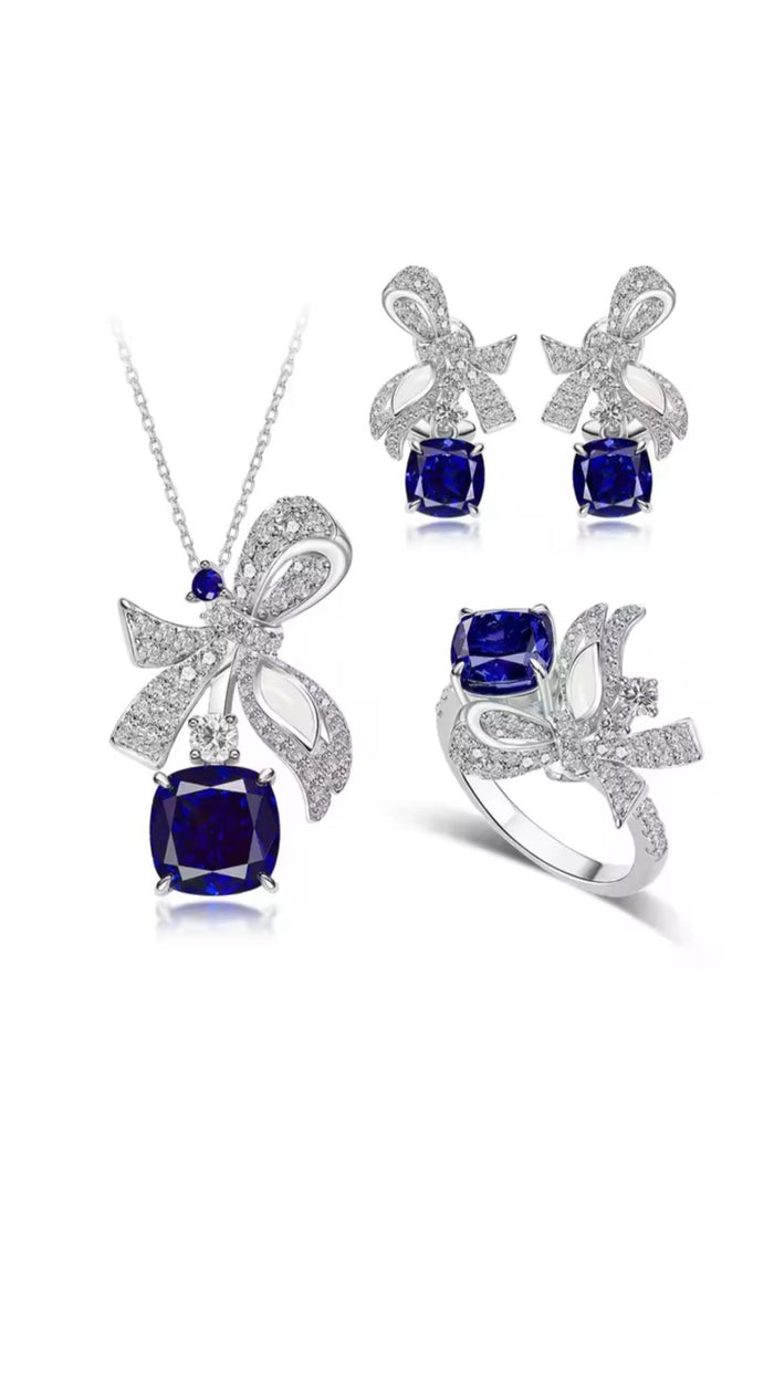 The Gem Vivi Blue Sapphire Drop Ribbon Accent Ring features a cushion-cut blue sapphire and silver bow accents with clear stones, crafted in 18k white gold. Pairs perfectly with matching necklace and earrings for a complete set.