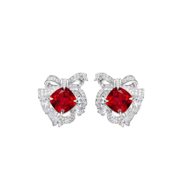 The Bloom Ribbon Stud Earrings by Gem Vivi are silver ribbon studs, each with a large cushion-cut ruby center surrounded by small clear gemstones.