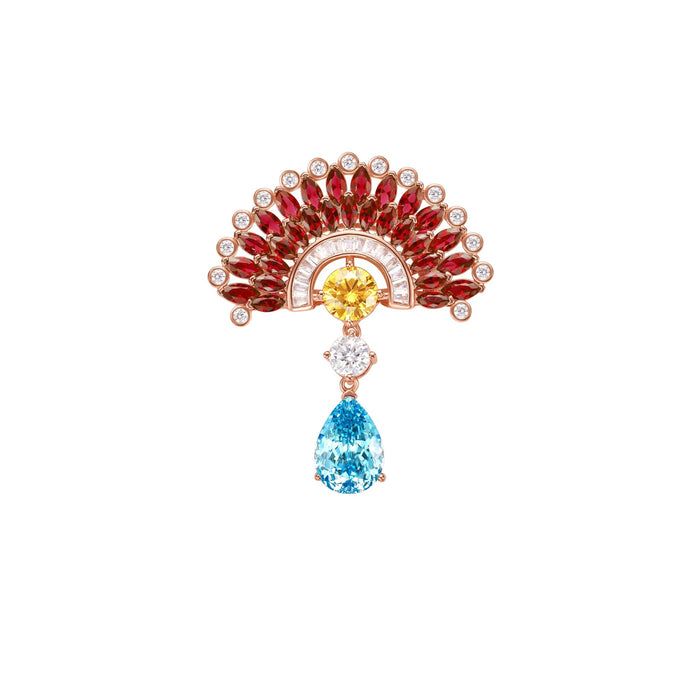 The Ruby Peacock Brooch by Gem Vivi showcases a fan-shaped peacock design with red and yellow gems, a central diamond, and a teardrop blue gem dangling below, all elegantly set in sterling silver.