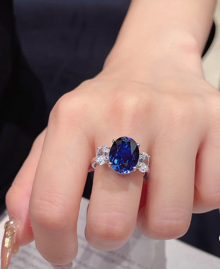 A hand displays the Gem Vivi Blue Sapphire Elegance Ring, featuring a large oval cut sapphire set in sterling silver and accented by two smaller clear stones.