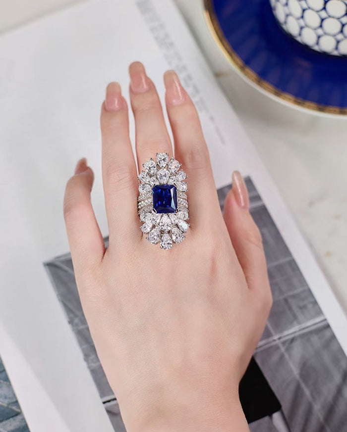 A hand wearing the Candela Radiance Regal Cluster Ring by Gem Vivi, featuring a rectangular blue gemstone encircled by clear stones, rests on an open magazine.
