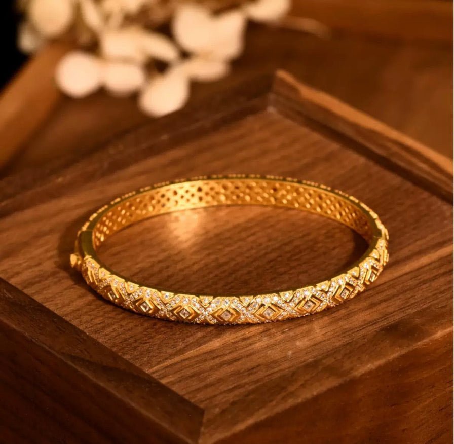 The Selena Lattice Bangle by Gem Vivi features intricate geometric patterns and embedded cubic zirconia stones in 18K yellow gold, elegantly displayed on a wooden surface.