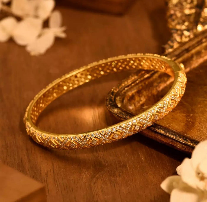 The Selena Lattice Bangle by Gem Vivi features intricate 18K yellow gold engravings, displayed on a wooden surface and accented with decorative white flowers.